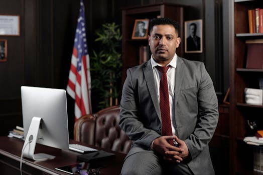 A professional businessman in a suit sitting in an office setting, with the American flag in the background.